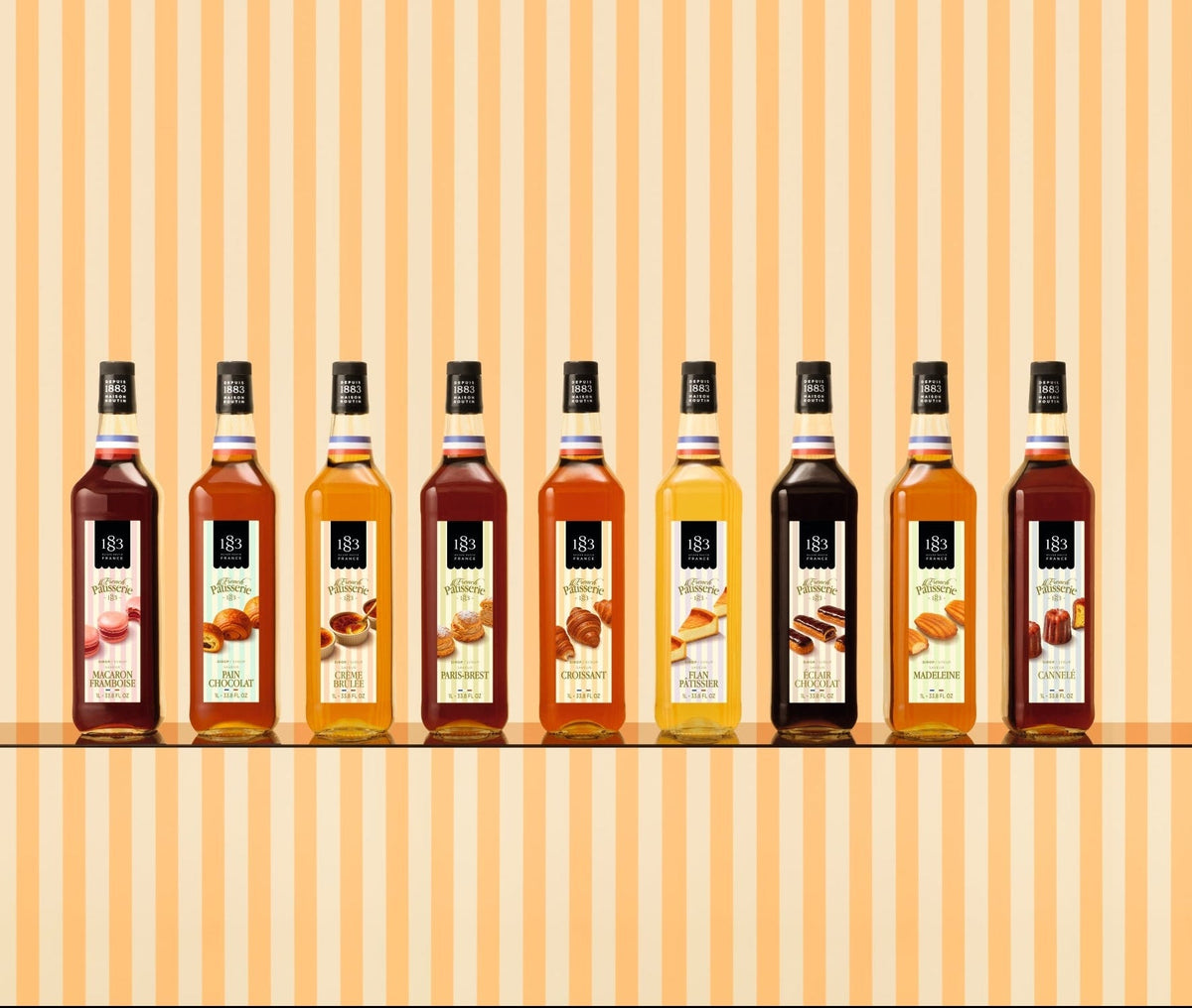 Eight bottles of syrup arranged in a row on a shelf against a warm beige and orange striped background. The bottles contain various colored syrups in shades of amber, brown, and golden yellow, each with a white label and black cap.