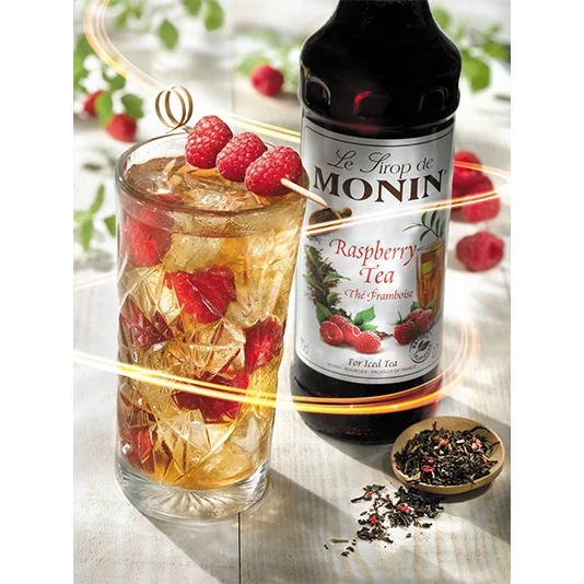 Glass of raspberry tea with ice and raspberries, Monin raspberry tea syrup bottle, and dried tea leaves on a white surface.
