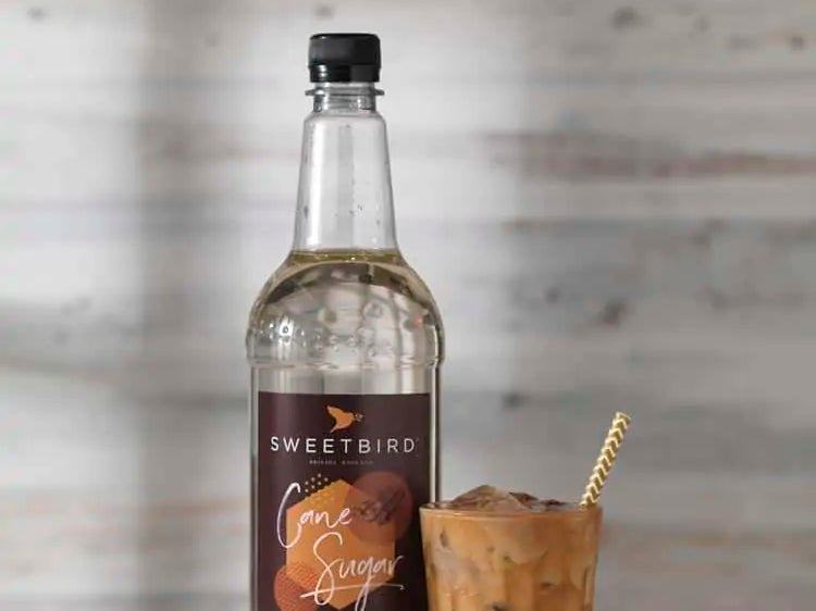 Bottle of Sweetbird Cane Syrup with a glass of iced coffee on a wooden surface.