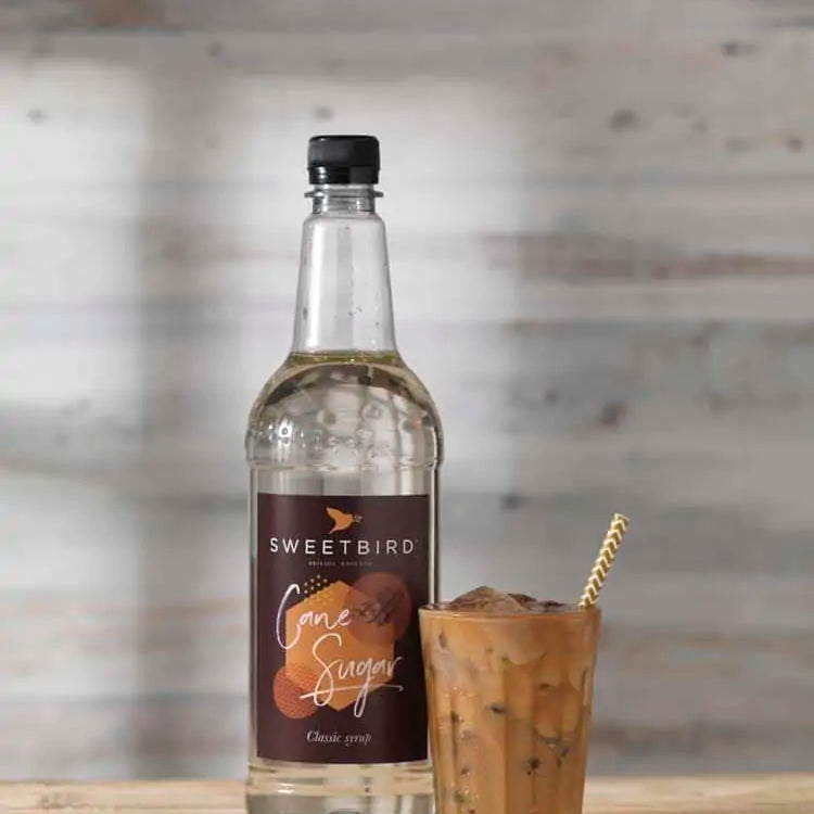 Bottle of Sweetbird Cane Syrup with a glass of iced coffee on a wooden surface.