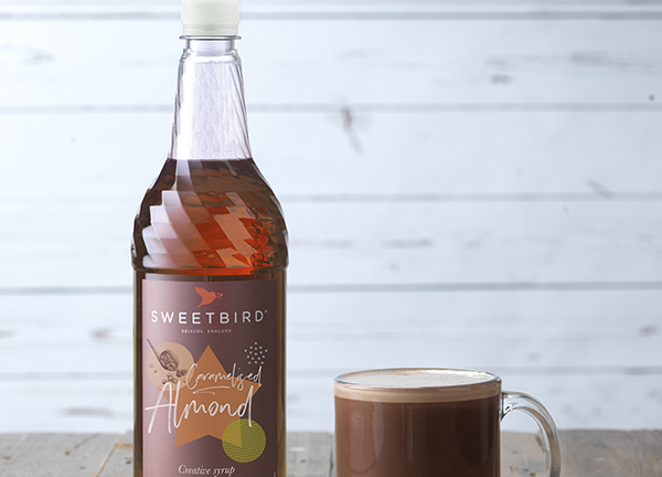 Bottle of Sweetbird Almond syrup next to a glass of hot chocolate on a wooden surface.