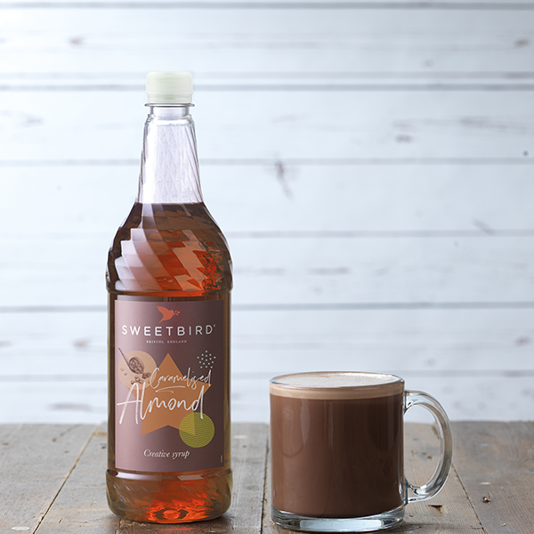 Bottle of Sweetbird Almond syrup next to a glass of hot chocolate on a wooden surface.