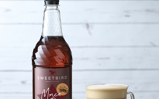 Bottle of Sweetbird Mac Pie syrup next to a cup of coffee on a wooden surface with a light gray background.