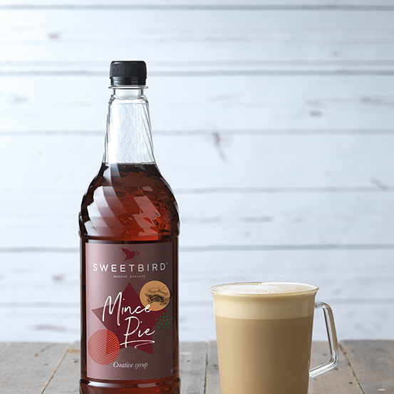 Bottle of Sweetbird Mac Pie syrup next to a cup of coffee on a wooden surface with a light gray background.