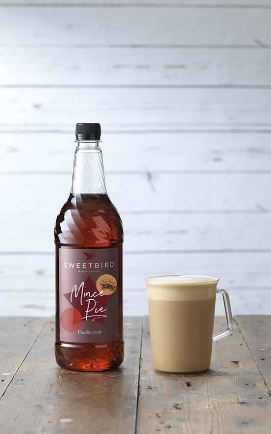 Bottle of Sweetbird Mac Pie syrup next to a cup of coffee on a wooden surface with a light gray background.