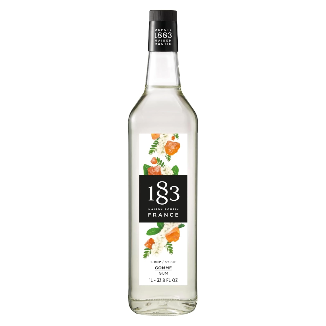 A bottle of 1883 Maison Matrio France Gomme Cane syrup, featuring a clear glass bottle with a white and black label decorated with orange and green floral illustrations, set against a plain white background.